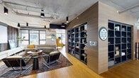 This thoughtfully designed multi-level space masterfully combines industrial elements with contemporary aesthetics. Exposed concrete ceilings with visible ductwork and pendant lighting contrast beautifully against warm oak hardwood flooring that flows throughout the property, creating an inviting urban atmosphere.

The main lounge area serves as a social hub, furnished with a curved sectional sofa, modern accent chairs in bold mustard yellow, and layered area rugs. A large built-in bookshelf partition clad in warm wood paneling doubles as a functional room divider and wayfinding feature, displaying books and decorative objects.

A dedicated media lounge offers a large projection screen, a plush sectional, and a marble-top coffee table, flanked by windows that bring in natural light. Adjacent to this, a bright corner seating area features floor-to-ceiling windows offering sweeping city street views, furnished with statement yellow wingback chairs and a marble round table.

A central mezzanine-style corridor with cable-rail balustrades overlooks a dramatic double-height lobby below, which is bathed in natural light from expansive glazed facades. The hallways are lined with exposed brick tile walls and globe pendant lighting, lending warmth and character. Glass-enclosed meeting or office spaces line the upper-level walkway, ensuring transparency and connectivity throughout.