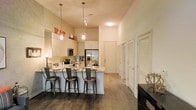 This single-floor residence showcases a thoughtfully designed open-concept layout that seamlessly blends the kitchen and living areas into one cohesive, functional space. The kitchen is a standout feature, boasting sleek, light-toned cabinetry that extends toward impressively high ceilings, maximizing the sense of vertical space throughout. A generous breakfast bar with stone countertops accommodates multiple bar stools, making it ideal for casual dining and entertaining. Stainless steel appliances add a contemporary touch to the kitchen, which is further elevated by stylish pendant lighting that hangs gracefully above the bar area. Rich, dark hardwood-style flooring runs throughout, providing a warm contrast to the bright, neutral-toned walls and cabinetry. Natural and ambient light fills the space, enhancing the airy, open feel. Multiple doors along the hallway suggest well-defined private rooms beyond the main living area. The overall ambiance is modern and inviting, with clean lines, quality finishes, and a layout designed to support both comfortable everyday living and effortless entertaining.