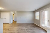 This single-floor unit presents a clean, modern aesthetic unified by wood-look plank flooring that runs consistently throughout each room. The interior is finished with soft gray walls and crisp white baseboards, creating a neutral palette well-suited to a variety of furnishing styles.

The living area is generously proportioned, receiving natural light through a window fitted with horizontal blinds. A glass-panel entry door adjoins a secondary window, further brightening the space and providing outdoor access. An open layout connects the living area to the kitchen, which is separated by a half-wall counter that functions as a pass-through or breakfast bar.

The kitchen is well-equipped with white cabinetry extending to the ceiling for ample storage, a white electric range, refrigerator, and under-cabinet range hood. A window above the sink area allows natural light into the cooking space.

The bedrooms are comfortably sized with the same consistent flooring and neutral gray walls. Each room features white doors and standard closet access. The bathroom includes a full tub and shower combination with a curtain rod, a vanity with a light-toned countertop, and wood-look flooring, maintaining the cohesive design found throughout the unit.