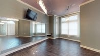 This single-floor studio space combines refined aesthetics with practical functionality, making it well-suited for fitness, dance, yoga, or wellness activities. The room is anchored by rich, dark hardwood flooring that extends throughout, providing a sleek and durable surface for a variety of uses. A full-length wall mirror spans a significant portion of one wall, creating depth and allowing users to monitor form and movement with ease. Soft sage-green walls are accented by crisp white crown molding and baseboards, lending the space a clean, polished character. Overhead, a row of glamorous crystal chandelier pendants serves as a striking design centerpiece, casting a warm and inviting glow across the room. Large, white-trimmed windows allow generous natural light to fill the space, complementing the bright and airy atmosphere. A wall-mounted flat-screen television is also present, enabling instructional or entertainment use during sessions. The combination of elegant finishes, reflective surfaces, and thoughtful lighting gives this studio a boutique feel that balances both sophistication and practicality.