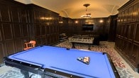 This elegant games room is defined by its striking architectural character, most notably a vaulted ceiling that adds volume and grandeur to the space. The walls are clad floor-to-ceiling in rich, dark wood paneling with detailed raised molding, lending a classic, club-like atmosphere that feels both refined and inviting.

At the center of the room, a full-size billiards table with a vivid blue felt surface serves as the primary focal point, making it ideal for entertaining. A foosball table positioned nearby adds a more casual gaming option, while a wall-mounted flat-screen television provides a media element suited for viewing during gatherings.

Underfoot, a bold animal-print carpet introduces a dynamic visual contrast against the dark paneling and anchors the entertainment area with a touch of contemporary style. Ambient lighting is provided by a central chandelier suspended from the apex of the vault, complemented by wall-mounted sconces that cast a warm, intimate glow throughout the room.

Together, these elements create a well-appointed games room that balances traditional architectural detail with practical recreational amenities.