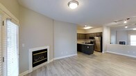 This Aspenwood apartment unit offers a thoughtfully designed single-floor layout that seamlessly blends comfort and modern style. The open-concept living area is anchored by a clean, white-framed fireplace with gold trim accents, providing a warm and inviting focal point for the space. Light wood-grain flooring runs throughout, lending a bright and cohesive feel to the entire unit.

The living area flows naturally into the kitchen, which is appointed with dark espresso cabinetry that creates a striking contrast against the neutral warm-toned walls. Stainless steel appliances, including a refrigerator, microwave, and range, lend a contemporary and functional edge to the kitchen. A central kitchen island with a built-in cooktop provides additional prep space and serves as a natural divider between the cooking and living areas. A sink with a window-adjacent position further enhances the kitchen's practical layout.

Overhead lighting is recessed and flush-mounted, providing even illumination throughout the unit. The neutral wall palette and clean white baseboards and trim contribute to a fresh, move-in-ready aesthetic. Overall, this unit at Aspenwood combines practical functionality with modern design elements, making it a comfortable and attractive living space.