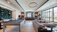 This expansive single-floor amenity space blends modern design with industrial-inspired accents to create a dynamic and inviting communal environment. Light hardwood flooring flows throughout, unifying the various activity zones beneath soaring ceilings punctuated by dramatic circular candle-style chandeliers and recessed lighting.

The game lounge anchors one end of the space, centering around a full-size pool table set against a striking wall of floor-to-ceiling dark teal shelving units framed in black steel. Built-in bench seating with cushions and side tables creates a comfortable spectator area. Exposed brick accent walls add warmth and texture throughout, complementing the otherwise crisp white palette.

A generous lounge area features a large sectional sofa, accent chairs in bold red, a flat-screen television, and large steel-framed windows that flood the room with natural light and offer views of a landscaped courtyard with palm trees and a pool beyond. The space opens further into a bright central hall with vaulted ceilings, pendant lighting, and an organic centerpiece installation.

A sleek bar and kitchen area features dual runs of dark cabinetry topped with expansive marble-veined countertops, gold hardware accents, and pendant lighting. A sink runs along the window-facing counter, bathed in natural light. High-top bar seating and intimate café-style tables near oversized windows complete this sophisticated and well-appointed communal retreat.