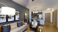 This single-floor residence showcases a thoughtfully designed open-concept layout that integrates the living, dining, and kitchen areas into one cohesive space. The living area is anchored by a neutral-toned sofa accented with navy and patterned throw pillows, complemented by a leather accent chair and a geometric-patterned area rug. A ceiling fan overhead adds both function and style. Bold charcoal-gray accent walls provide a striking contrast against lighter surrounding walls, adding depth and visual interest throughout.

The kitchen is a standout feature, boasting dark cabinetry, stainless steel appliances, and a peninsula with bar-height seating illuminated by sleek pendant lighting. Light wood-tone flooring flows consistently throughout, unifying the space and enhancing its warmth. The open sightlines between zones create an airy, connected feel ideal for both everyday living and entertaining. Contemporary framed artwork adorns the walls, reinforcing the modern aesthetic. Overall, the space balances bold design choices with functional, well-appointed details.