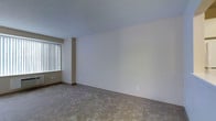 This single-floor residential space presents a clean, minimalist aesthetic defined by neutral color tones and straightforward design. The interior is finished with wall-to-wall plush carpeting in a warm beige hue, providing a soft and comfortable underfoot experience throughout. The walls are painted in a smooth, light neutral tone that lends the space a bright and airy feel, complemented by a flat white ceiling that enhances the sense of openness. Natural light enters through a large window dressed with vertical blinds, allowing for flexible light control while maintaining privacy. The window treatment and architectural column detail near the window add subtle visual interest to an otherwise open and unobstructed layout. Electrical outlets are positioned conveniently along the walls, reflecting a practical and functional design approach. The overall ambiance of the space is calm and understated, making it a versatile canvas well-suited for a variety of interior arrangements and personal styles. The single-floor layout ensures easy accessibility and seamless flow throughout the space.