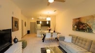 This single-floor residence showcases an open-concept layout that seamlessly connects the living, dining, and kitchen areas into one cohesive space. The living area is anchored by plush carpeting and features a ceiling fan overhead, contributing to a comfortable atmosphere. A glass-topped coffee table and a contemporary sofa with accent pillows complete the lounge area, offering a relaxed yet polished aesthetic.

Adjacent to the living space, the dining area accommodates a modern rectangular table with a bold geometric base, paired with sleek white chairs. Above the dining table, a rectangular pendant light fixture provides focused illumination, adding a stylish focal point to the room.

The kitchen, visible in the background, features dark cabinetry contrasted against light countertops, with stainless steel appliances including a refrigerator and microwave. A kitchen island or breakfast bar with bar seating creates an additional casual dining option and enhances functionality.

Warm ambient lighting from multiple sources, including pendant lights and ceiling fixtures, gives the entire space an inviting glow. Neutral wall tones and light-colored flooring contribute to a bright, airy feel throughout.