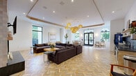 Located in North Miami Beach, FL, this single-floor combined living and dining space offers a thoughtfully designed interior that balances modern elegance with warm, coastal-inspired elements. The standout herringbone-patterned tile flooring runs throughout the entire space, lending a sophisticated visual texture that anchors the open layout.

The living area is centered around a striking stone-clad fireplace with a mounted television above, flanked by a generous sectional sofa and accent seating. Recessed ceiling lighting and a tray ceiling detail enhance the sense of height and architectural definition. A compact kitchenette with dark tile backsplash and built-in appliances is neatly integrated into the space.

The dining area is framed by a series of arched, black-framed windows that flood the room with natural light and offer views of lush tropical greenery. Glass-top round tables paired with woven rattan chairs create a relaxed yet refined dining atmosphere. A cluster of warm-toned woven pendant lights anchors the transition between living and dining zones, adding an inviting glow.

Oversized black-framed double doors provide direct access to the outdoors, reinforcing the connection to the surrounding landscape. The overall ambiance is bright, airy, and polished, making this a versatile and welcoming communal space.