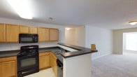 Located at 215 William Penn Plz in Durham, North Carolina, this single-floor residence offers a practical and well-organized layout designed for comfortable everyday living. The kitchen serves as a central feature of the space, outfitted with warm oak cabinetry that provides ample storage, dark laminate countertops, and a full suite of black appliances including a wall oven, built-in microwave, and dishwasher. A double-basin sink sits beneath the counter, and the kitchen flows naturally into the living area by way of a peninsula-style breakfast bar — ideal for casual dining or entertaining. The open-concept design between the kitchen and living room enhances the sense of space, with neutral-toned carpeting in the living area creating a soft, cohesive aesthetic. A textured ceiling runs throughout, and the light, neutral wall colors contribute to a bright and airy feel. Flush-mounted lighting keeps the space well-lit, while multiple electrical outlets and HVAC vents reflect thoughtful utility planning throughout. Overall, the layout balances functionality with an open, approachable design that suits a range of lifestyles.
