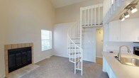 This thoughtfully designed space makes an immediate impression with its dramatically high vaulted ceilings, which create an airy, open atmosphere throughout the main living area. The standout architectural feature is a white spiral staircase with carpeted treads and classic spindle railings, elegantly connecting the main floor to an upper loft area above. The loft overlooks the living space below, adding a sense of vertical dimension and versatility to the layout.

The main living area is finished with neutral wall-to-wall carpeting that provides a warm, comfortable feel underfoot, while the adjacent area transitions to hard surface flooring, offering practical contrast. A window with horizontal blinds allows natural light to filter into the space, brightening the neutral, softly toned walls.

A fireplace is visible along one wall, contributing a cozy focal point to the living area. The loft level above provides additional usable space, accessible exclusively via the spiral staircase, and is partially enclosed with a railing for safety. The overall ambiance is calm and neutral, offering a versatile canvas adaptable to a variety of interior styles and uses.