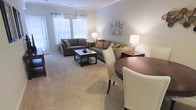 This single-floor residence features an open-concept layout that seamlessly connects the living and dining areas, creating a cohesive and functional flow throughout the space. The living area is anchored by two plush, charcoal-gray sofas arranged around a dark wood coffee table, providing a comfortable and inviting seating arrangement. Accent pillows in warm earth tones add visual interest against the neutral upholstery. Soft, neutral-toned carpeting extends throughout the space, contributing to a warm and comfortable atmosphere. The walls are painted in a calming light gray-blue hue, complementing the overall neutral palette. Crown molding along the ceiling adds a refined architectural detail. Natural light filters generously into the living area through large windows dressed with sheer white curtains, creating a bright and airy ambiance. Table lamps on either side of the seating area provide warm supplemental lighting. The dining area, positioned adjacent to the living space, features a round dark wood table paired with elegant cream upholstered chairs. A colorful abstract artwork on the wall serves as a subtle decorative focal point, adding a touch of personality to the otherwise understated interior.