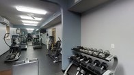 This fitness center is a single-floor facility designed to provide a thorough and versatile workout experience. The space is organized into distinct exercise zones, making efficient use of its layout. Upon entering, a dedicated free weights area greets users with a full rack of dumbbells in varying sizes, neatly arranged and easily accessible for strength training exercises.

Beyond the free weights section, the main floor opens up to a lineup of cardio equipment, including treadmills and an elliptical cross-trainer, positioned along the length of the room to allow for comfortable, unobstructed use. A ceiling fan ensures air circulation throughout the cardio area, contributing to a more comfortable workout environment.

The facility features durable rubber flooring suited for heavy equipment and high-impact activity, while the walls are finished in a cool blue-gray tone that lends a focused, energetic atmosphere. Overhead fluorescent lighting ensures the entire space is well-lit at all times. A wall-mounted television is visible near the cardio section, offering entertainment during longer sessions. The space concludes with a doorway at the far end, suggesting additional utility or access areas. Overall, this fitness center is thoughtfully arranged to accommodate both cardio and strength training needs efficiently.