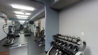 This fitness center is a single-floor facility designed to provide a thorough and versatile workout experience. The space is organized into distinct exercise zones, making efficient use of its layout. Upon entering, a dedicated free weights area greets users with a full rack of dumbbells in varying sizes, neatly arranged and easily accessible for strength training exercises.

Beyond the free weights section, the main floor opens up to a lineup of cardio equipment, including treadmills and an elliptical cross-trainer, positioned along the length of the room to allow for comfortable, unobstructed use. A ceiling fan ensures air circulation throughout the cardio area, contributing to a more comfortable workout environment.

The facility features durable rubber flooring suited for heavy equipment and high-impact activity, while the walls are finished in a cool blue-gray tone that lends a focused, energetic atmosphere. Overhead fluorescent lighting ensures the entire space is well-lit at all times. A wall-mounted television is visible near the cardio section, offering entertainment during longer sessions. The space concludes with a doorway at the far end, suggesting additional utility or access areas. Overall, this fitness center is thoughtfully arranged to accommodate both cardio and strength training needs efficiently.