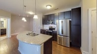 This single-floor residence offers a thoughtfully designed open-concept layout that seamlessly connects the kitchen, dining, and living areas. The kitchen is a standout feature, equipped with rich dark cabinetry, stainless steel appliances including a refrigerator, range, and built-in microwave, and a granite-topped peninsula with an undermount sink and pendant lighting above. The living area extends naturally from the kitchen, highlighted by hardwood-style flooring, track lighting, and a glass-paneled door that brings in generous natural light and provides access to an outdoor area. The bedroom is carpeted for comfort, fitted with a ceiling fan with light, and benefits from natural light through a window with blinds. The primary bathroom features a dark wood vanity with granite countertops, a tub-shower combination with white tile surround, and brushed nickel fixtures. A second full bathroom mirrors the quality, offering a matching granite-topped vanity and a tub-shower unit. Throughout the space, warm beige walls, white trim, and consistent hardwood-style flooring in the main living areas create a cohesive and inviting atmosphere.
