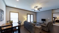 This single-floor space presents an open-concept layout that seamlessly connects the living, dining, and kitchen areas into one cohesive environment. The living room is generously sized, accommodating plush, dark upholstered sofas arranged around a glass-top coffee table, with a ceiling fan with integrated lighting fixture overhead and recessed lighting throughout. Crown molding trims the neutral-toned walls, adding a refined architectural detail to the space.

Dark hardwood flooring runs through the main living and dining areas, providing a warm contrast to the light-colored walls and ceiling. Multiple windows fitted with horizontal blinds allow natural light to filter throughout the space. The dining area features a dark wood table and matching chairs, positioned near the windows.

The kitchen is defined by a raised breakfast bar peninsula, dark cabinetry, and stainless steel appliances including a refrigerator and microwave. Pendant lights hang above the kitchen area, complementing the recessed lighting above. Neutral-toned doors lead to additional rooms, maintaining a consistent, understated color palette throughout the space.