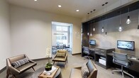 This single-floor space showcases a sleek, contemporary design that seamlessly blends comfort with functionality. The main area features a welcoming lounge with mid-century modern seating, including leather chairs with striped accent pillows, arranged around a round coffee table adorned with a white orchid centerpiece, adding a refined, natural touch.

A standout feature is the built-in dark-toned workstation alcove, illuminated by a series of stylish Edison-style pendant lights at varying heights. The workstation is equipped with a desktop computer, built-in shelving, and ample drawer storage, making it an ideal space for focused work.

Adjacent to the lounge, a glass-walled conference room offers a semi-private meeting area furnished with a long table and ergonomic mesh chairs. A large window floods the conference room with natural light, creating an open, airy atmosphere.

The overall design incorporates neutral tones — warm beige walls, light carpeting, and dark cabinetry accents — creating a cohesive and professional ambiance. Recessed ceiling lighting throughout ensures consistent illumination, complementing the space's polished, modern aesthetic.