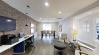 This thoughtfully designed single-floor space blends modern aesthetics with functional versatility. A striking exposed brick accent wall runs along one side of the room, adding warmth and industrial character that contrasts beautifully with the clean white walls and crisp crown molding throughout. Dark hardwood floors anchor the space, lending a sense of richness and continuity from one end to the other.

The layout is organized into distinct zones, each serving a unique purpose. A dedicated workstation area near the brick wall features a sleek desk, an ergonomic task chair, and a pendant light overhead, making it well-suited for focused work. Adjacent to this, a comfortable lounge area with an upholstered armchair, a tufted round ottoman, and a sculptural side lamp provides an inviting spot for casual conversation or relaxation.

Toward the far end of the room, a high-top communal table surrounded by tall chairs sits beneath a modern linear chandelier, positioned in front of a bright window that draws in natural light. Recessed ceiling lighting throughout ensures the entire space remains well-lit and welcoming. The overall ambiance is polished yet approachable, balancing contemporary design elements with practical, multi-use functionality.