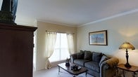 This single-floor space features a well-appointed living room that blends classic design elements with a warm, inviting ambiance. The room is finished with crown molding along the ceiling perimeter, adding a refined architectural detail that enhances the overall elegance of the space. Neutral, warm-toned walls create a calm backdrop throughout.

The living area is centered around a plush, grey upholstered sofa adorned with decorative pillows, paired with a traditional-style wooden coffee table featuring ornate detailing. A framed artwork piece hangs on the wall, contributing to the room's curated aesthetic. A dark wood armoire or cabinet is partially visible near the entrance, offering both storage and a grounding visual element.

Natural light fills the room through a large window dressed with sheer, cream-colored tab-top curtains that soften the incoming light while maintaining an airy feel. Light-toned carpeting adds warmth underfoot and complements the room's overall neutral palette. The space presents a comfortable and classically styled environment suitable for everyday living and relaxing.