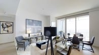 This refined single-floor residence exudes modern elegance through its thoughtfully designed open-plan layout. The living and dining area form a seamlessly connected space, bathed in natural light streaming through floor-to-ceiling glass doors that lead to a private balcony. The balcony extends the living experience outdoors, offering an airy retreat framed by a glass balustrade.

The living area is anchored by a plush sofa ensemble in neutral tones, complemented by a sleek console table adorned with decorative accents and a striking large-scale abstract artwork in muted blues and greys. A bold tripod floor lamp adds a sculptural focal point to the room. The dining area features a transparent glass-top table surrounded by contemporary dark chairs, creating a chic and practical dining setting.

Throughout, the interior palette is kept crisp and cohesive — white walls and light-toned flooring reflect the abundant natural light, enhancing the sense of openness and scale. Carefully curated accessories, including greenery and decorative objects, add warmth and character. The overall ambiance is one of understated luxury, with clean lines, high-quality finishes, and a calm, sophisticated atmosphere that is both welcoming and stylish.