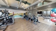 This fitness center is thoughtfully designed to accommodate a wide range of workout styles within a single, open-plan space. The architecture is immediately striking, with vaulted ceilings supported by exposed structural beams that lend an airy, industrial character to the room. Large windows along multiple walls flood the interior with natural light while offering views of the surrounding environment, creating an energizing atmosphere.

The space is organized into distinct functional zones, subtly separated by elegant metallic chain curtains that allow light to pass through while defining each area. The cardio zone features a row of treadmills, ellipticals, and a stair climber positioned beneath the expansive window wall. Nearby, a dedicated cycling area hosts multiple spin bikes alongside a wall-mounted television, ideal for group or solo ride sessions.

A mirrored studio area with a smooth wood-tone floor provides ample room for stretching, functional training, and group classes. This zone is equipped with stability balls, TRX suspension trainers, and step platforms. A scenic landscape mural adds a dynamic visual element to one wall. The flooring transitions between wood-tone finishes across zones, and pendant and track lighting complement the natural light throughout.
