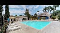 This inviting outdoor recreational facility centers around a generously sized swimming pool with clearly marked lap lanes, making it suitable for both leisure swimming and structured exercise. The crystal-clear, turquoise water is complemented by a wide surrounding concrete sundeck that provides ample space for relaxation and movement.

The pool area is well-equipped with white lounge chairs positioned along the poolside, offering comfortable spots for sunbathing. Shaded seating areas with patio umbrellas and outdoor furniture are situated to the sides, providing a respite from the sun for those who prefer a cooler vantage point.

Framing the space are one-story stucco buildings with sliding glass doors that open directly onto the pool deck, suggesting accessible amenity or facility rooms. Decorative potted plants and shrubs are thoughtfully placed around the perimeter, adding a touch of greenery and visual interest.

The landscaping features a mix of mature shade trees and tall palm trees that rise above the roofline, creating a lush, California-inspired ambiance. The open sky above allows for abundant natural light throughout the day, enhancing the resort-like atmosphere of this community outdoor space.