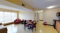 This single-floor space offers a welcoming and functional layout designed for comfortable everyday living. The open living and dining area serves as the heart of the unit, featuring warm yellow walls that create a bright and inviting atmosphere. The dining area is centered around a round dark-wood table accompanied by cushioned chairs with red upholstery, providing a cozy setting for meals. A matching armchair with red fabric accents anchors the adjacent sitting area, complemented by a low wooden shelving unit.

The flooring throughout is a light-toned vinyl or linoleum material that is easy to maintain and contributes to the clean, practical aesthetic of the space. Natural light filters in through a window in the living area, enhancing the airy feel of the room. Overhead ventilation grilles are integrated into the ceiling, ensuring climate control throughout.

A long hallway extends from the main living area, lined with doors leading to additional rooms, and illuminated by ceiling-mounted lighting fixtures. The hallway also features a utility panel mounted on the wall, indicating functional infrastructure within the space. Overall, the layout is straightforward and efficient, making it well-suited for residential use in a communal or assisted-living setting.