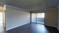 This single-story space presents a clean, functional layout highlighted by several notable design elements. The main living area features dark hardwood-style flooring that runs throughout, providing a sleek, modern contrast against the neutral-toned walls. Crown molding lines the upper perimeter of the room, adding a subtle architectural detail that elevates the overall finish of the space.

The ceiling carries a textured popcorn finish, a classic feature that adds dimension overhead. A generous sliding glass door dominates one wall, dressed with vertical blinds that allow for adjustable light control. When open, the blinds reveal a view of lush greenery outside, inviting an abundance of natural light into the interior and creating a seamless visual connection to the outdoors.

The neutral wall palette keeps the space feeling open and versatile, making it easy to personalize with various furnishing styles. The overall ambiance is bright and airy during the day, thanks to the expansive glazed door, while the dark flooring grounds the room with a sense of warmth and depth. The space offers a straightforward, well-maintained environment suited for comfortable everyday living.