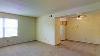 This single-floor space features an open layout that connects the main living area to the kitchen, creating a functional and cohesive floor plan. The living area is finished with wall-to-wall carpeting in a neutral tone, complemented by light-colored walls that lend a clean, understated aesthetic. A ceiling vent ensures year-round climate comfort throughout the space.

The kitchen is accessible through an open passageway, offering a seamless transition between living and cooking areas. It features tile flooring, warm-toned wood cabinetry mounted above, and a white refrigerator. A ceiling fan is installed in the kitchen area, providing additional air circulation. The open-concept connection between the two spaces allows for easy interaction and a sense of continuity throughout the home.

Overall, the space maintains a simple, functional design with neutral finishes throughout. The consistent color palette of cream and beige tones across both rooms contributes to a cohesive and airy atmosphere, making it a practical and adaptable living environment.