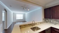 This single-floor space showcases an open-concept layout that seamlessly connects the kitchen and living area. The kitchen is appointed with light-toned countertops, a stainless steel undermount sink, and dark cabinetry, complemented by a decorative mosaic tile backsplash featuring a blend of earth tones and neutrals. A breakfast bar-style peninsula serves as a natural divider between the kitchen and the adjoining living space, enhancing the open flow of the layout. Track lighting is mounted overhead, providing directional illumination throughout the transition area, while crown molding runs along the ceiling, adding a refined architectural detail. The living area features dark hardwood flooring and is bathed in natural light from a large window dressed with horizontal blinds. Neutral gray walls throughout the space create a clean, cohesive aesthetic. The open-concept design and thoughtful layout make this space well-suited for both everyday living and entertaining.