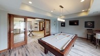 This single-floor game and recreation space is thoughtfully designed for both leisure and socializing. The centerpiece of the room is a full-size pool table with a light grey felt surface, rich wood rail trim, and a modern rectangular pendant light suspended above it. Recessed lighting complements the pendant fixture, ensuring the space is well-lit throughout.

The flooring features distressed wood-look planks in warm, varied tones, adding texture and visual interest underfoot. Neutral grey walls are accented by warm wood baseboards and window trim, creating a cohesive and polished aesthetic. A wall-mounted flat-screen television is positioned for easy viewing, while billiards-themed wall art adds character to the space.

A row of large, dark wood-trimmed windows along one wall fills the room with natural light and offers views of an outdoor patio and surrounding greenery. A glass-paneled sliding door connects the game room to adjacent spaces, including what appears to be a lounge and kitchen area, providing a sense of openness and flow. The overall ambiance is casual yet refined, making this a versatile space well-suited for recreation and entertaining.