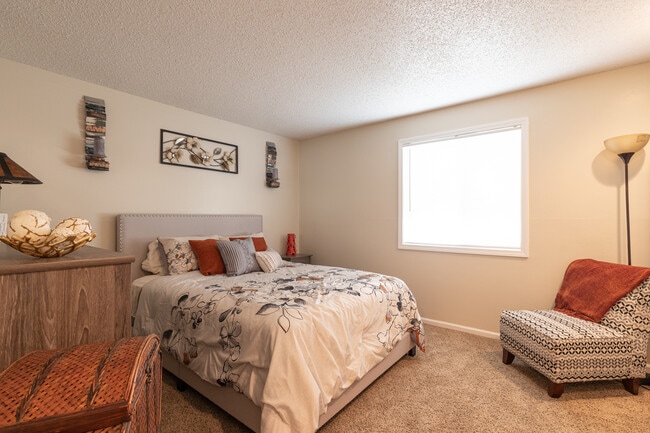 A spacious bedroom with a beige padded headboard, floral black-stitch bedding, a natural wood dresser, a cozy orange lounge chair, and a window with a scenic view. - Wilshire Apartments