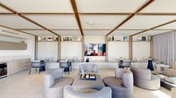 This elegantly designed single-floor lounge offers a refined open-plan layout that masterfully blends interior comfort with outdoor living. The interior is distinguished by a striking coffered ceiling with warm-toned exposed wood beams set against a crisp white backdrop, creating a sense of architectural depth and character. Polished terrazzo-style flooring runs throughout, lending a clean, upscale finish to the space.

The main lounge area features a generous sectional sofa in neutral tones, rounded upholstered accent chairs, and a sleek central coffee table, all oriented around a large wall-mounted flat-screen display. Built-in banquette seating alcoves line one wall, each framed by light wood shelving displaying curated décor, with individual bistro-style tables and woven accent chairs providing intimate seating nooks.

Floor-to-ceiling sliding glass panels open fully to a spacious outdoor terrace, blurring the boundary between inside and out. The terrace is equipped with modular outdoor sofas, lounge chairs, and shaded seating areas beneath a pergola-style overhead structure. Lush palm trees and panoramic skyline views form the backdrop, bathing the entire space in abundant natural light. A compact kitchenette is also visible, adding functional utility to this polished, resort-inspired environment.