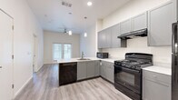 This well-designed one-bedroom, one-bathroom unit in Austin, TX offers a thoughtfully laid-out single-level floor plan with modern finishes throughout. The open-concept kitchen serves as the heart of the space, featuring gray shaker-style cabinetry, light stone countertops, a peninsula with an integrated sink, and black appliances including a gas range, dishwasher, refrigerator, and microwave. Pendant lighting hangs above the peninsula, adding a contemporary touch, while light wood-look flooring extends across the main living area. The kitchen opens naturally to the living area, which benefits from natural light through a set of windows framed with blinds and a ceiling fan overhead. The bedroom is carpeted for comfort and includes a window that brings in additional natural light, along with access to a dedicated closet. The bathroom is spacious and thoughtfully designed with accessibility in mind, featuring grab bars, a combination tub and shower with a handheld showerhead, a wide vanity with a stone countertop, gray cabinetry, and a large mirror. The consistent use of light tones, clean lines, and modern fixtures throughout creates a cohesive and functional living environment.
