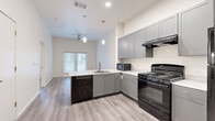 This well-designed one-bedroom, one-bathroom unit in Austin, TX offers a thoughtfully laid-out single-level floor plan with modern finishes throughout. The open-concept kitchen serves as the heart of the space, featuring gray shaker-style cabinetry, light stone countertops, a peninsula with an integrated sink, and black appliances including a gas range, dishwasher, refrigerator, and microwave. Pendant lighting hangs above the peninsula, adding a contemporary touch, while light wood-look flooring extends across the main living area. The kitchen opens naturally to the living area, which benefits from natural light through a set of windows framed with blinds and a ceiling fan overhead. The bedroom is carpeted for comfort and includes a window that brings in additional natural light, along with access to a dedicated closet. The bathroom is spacious and thoughtfully designed with accessibility in mind, featuring grab bars, a combination tub and shower with a handheld showerhead, a wide vanity with a stone countertop, gray cabinetry, and a large mirror. The consistent use of light tones, clean lines, and modern fixtures throughout creates a cohesive and functional living environment.