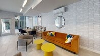 Located at 417 Centinela Ave in Inglewood, California, this co-working lounge is a thoughtfully designed single-floor workspace that balances productivity and comfort. The interior is defined by textured white brick walls that serve as a neutral backdrop for a curated collection of bold, contemporary artwork, lending the space a creative and inspiring atmosphere. Large-format light tile flooring runs throughout, contributing to the bright, open feel of the room. Floor-to-ceiling glass windows and sliding doors line one side of the lounge, flooding the interior with natural light and offering direct views of and access to an outdoor courtyard area. A long, solid wood communal worktable anchors the workspace, surrounded by ergonomic rolling chairs in deep navy, ideal for focused individual work or collaboration. Adjacent to the workstations, a lounge area provides a more relaxed setting, featuring a plush amber velvet sofa accented with colorful throw pillows, paired with vibrant yellow accent tables and additional upholstered seating. Recessed and linear LED ceiling lighting supplements the natural light, ensuring a well-lit environment throughout the day. A wall-mounted mini-split climate control unit provides personalized comfort. The overall ambiance is modern, energetic, and welcoming—well-suited for a dynamic co-working community.