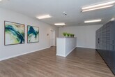 Located at 117 Teakwood Dr in Greensboro, NC, this single-floor commercial space serves as a well-appointed leasing or community office. The interior is finished with warm wood-look luxury vinyl plank flooring that runs consistently throughout, complemented by crisp white walls and clean white baseboards that give the space a bright, modern feel.

The main reception area is anchored by a prominent front counter with a light stone-top surface, accented by potted greenery that adds a welcoming touch. Two large abstract artworks in teal and blue tones hang on the wall, contributing a polished, professional aesthetic. Recessed and flush-mounted lighting fixtures provide consistent, even illumination across the space.

A dedicated package management area houses a large bank of smart package lockers, offering convenient and secure parcel retrieval. Adjacent to the lockers, a glass-panel exterior door opens to a greenery-lined outdoor area, allowing natural light to filter into the room. A utility table provides additional workspace for sorting and staging packages and deliveries.

The hallway connecting the rooms is clean and functional, featuring a security camera and HVAC vents, reinforcing the practical design of the space. Throughout, the layout prioritizes ease of navigation and operational efficiency, making this a functional and attractive facility for day-to-day office and resident services use.