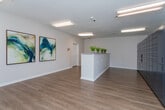 Located at 117 Teakwood Dr in Greensboro, NC, this single-floor commercial space serves as a well-appointed leasing or community office. The interior is finished with warm wood-look luxury vinyl plank flooring that runs consistently throughout, complemented by crisp white walls and clean white baseboards that give the space a bright, modern feel.

The main reception area is anchored by a prominent front counter with a light stone-top surface, accented by potted greenery that adds a welcoming touch. Two large abstract artworks in teal and blue tones hang on the wall, contributing a polished, professional aesthetic. Recessed and flush-mounted lighting fixtures provide consistent, even illumination across the space.

A dedicated package management area houses a large bank of smart package lockers, offering convenient and secure parcel retrieval. Adjacent to the lockers, a glass-panel exterior door opens to a greenery-lined outdoor area, allowing natural light to filter into the room. A utility table provides additional workspace for sorting and staging packages and deliveries.

The hallway connecting the rooms is clean and functional, featuring a security camera and HVAC vents, reinforcing the practical design of the space. Throughout, the layout prioritizes ease of navigation and operational efficiency, making this a functional and attractive facility for day-to-day office and resident services use.