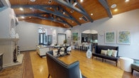 This single-floor lobby makes an immediate impression with its dramatic vaulted ceiling adorned with dark-painted exposed trusses and warm wood paneling, creating a bold architectural statement that defines the entire space. Rich hardwood floors extend throughout, complementing the clean white walls and dark-toned accents found in the built-in cabinetry and trim details.

The open layout is organized into distinct yet flowing zones. A central lounge area features a stylish arrangement of white upholstered sofas, leather accent chairs with nailhead trim, and a bold geometric console table, all anchored by a patterned area rug. A stone fireplace flanked by dark built-in bookshelves and display niches serves as a striking focal point along one wall, adding warmth and sophistication.

Adjacent to the main lounge, a bar-height counter with white stools provides a casual gathering area, while a kitchen-style space with dark cabinetry is accessible through an arched passageway. Colorful artwork punctuates the walls throughout.

At the far end, a windowed sitting area with floor-to-ceiling windows and transoms floods the space with natural light and offers views of lush greenery and a pool area beyond. This sunlit nook, furnished with tufted wingback chairs and sleek benches, provides a serene retreat within the larger lobby.