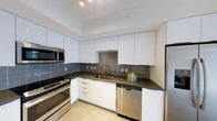 This single-floor space showcases a well-appointed kitchen with a contemporary aesthetic. The kitchen is designed with clean lines and a neutral color palette, featuring flat-front white cabinetry that extends to the ceiling, maximizing storage capacity. Dark charcoal countertops provide a striking contrast against the white cabinets, while a matching large-format tile backsplash adds texture and visual depth to the space.

The kitchen is fully equipped with a suite of stainless steel appliances, including a slide-in range, built-in microwave, dishwasher, and a refrigerator. The undermount sink is positioned at the base of the backsplash, complemented by a modern arc-neck faucet. Recessed track lighting installed in the white ceiling ensures even, practical illumination throughout the workspace.

The L-shaped layout of the kitchen promotes efficient workflow, with ample counter space along both walls. Light-toned flooring ties the overall design together, reinforcing the bright and modern character of the space. The overall ambiance is clean, functional, and polished, reflecting a contemporary design sensibility.