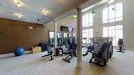 This fitness center offers a thoughtfully designed workout environment that balances functionality with an inviting atmosphere. The space features soaring ceilings with large, arched windows along one wall that bathe the room in abundant natural light, creating an open and energizing ambiance. Elegant pendant lighting and flush-mount fixtures complement the natural illumination throughout. The warm, two-tone color scheme — with rich brown accent walls and neutral upper walls — adds a polished aesthetic to the space. Sturdy white structural columns define the layout while maintaining an open feel. The workout floor is finished with light wood-style flooring, providing a clean and durable surface underfoot. A comprehensive selection of strength training equipment is centrally arranged, including cable machines, adjustable benches, and a Rogue-branded weight box, catering to a variety of training needs. Along the windowed wall, a dedicated cardio zone houses treadmills and additional machines. Full-length mirrors line one wall, enhancing the sense of space and allowing for proper form checks. Dual flat-screen TVs are mounted for entertainment during workouts. This facility is well-suited for a complete and effective fitness routine.