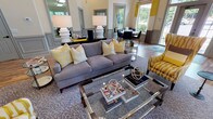 This clubhouse offers a well-appointed, single-floor community gathering space defined by a cohesive and contemporary design aesthetic. The interior is anchored by a bold yellow and gray color palette, carried throughout via upholstered furnishings, decorative accents, and striking striped drapery panels.

The main lounge area features soaring vaulted ceilings with a beadboard finish and an impressive arched window ensemble that draws in generous natural light and provides views of the surrounding greenery. The space is furnished with a plush gray sectional sofa accented by yellow throw pillows, complemented by a chrome-framed glass coffee table and a pair of yellow-and-white wingback chairs. Elegant wainscoting lines the walls throughout, adding a refined architectural touch.

Adjacent to the lounge, a dining nook with bistro-style tables and chairs provides additional seating for residents. Bold yellow glass panel artwork adorns the walls, reinforcing the vibrant design scheme. Hardwood-style flooring flows seamlessly across the entire space, enhancing the open and cohesive layout.

A dedicated fitness center is accessible directly from the main lounge, featuring an array of cardio and exercise equipment visible through interior windows. Large exterior windows and glass doors throughout the clubhouse ensure the space remains bright and connected to the outdoors.