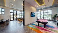 This contemporary amenity lounge is a dynamic, single-floor space thoughtfully designed to encourage socializing and recreation. The open layout is anchored by a bold, colorful billiards table set atop a vivid geometric area rug, positioned beneath a tray ceiling with warm amber accent lighting that adds depth and energy to the room. A wall-mounted flat-screen TV and light wood panel accent wall serve as a central focal point, complementing the polished large-format tile flooring throughout.

Floor-to-ceiling black-framed windows flood the space with natural light and offer sweeping views of a landscaped courtyard surrounded by a residential community. Multiple distinct seating areas create inviting zones within the open plan — including a cozy lounge corner with tufted blue leather armchairs arranged around a coffee table beneath oversized windows, and a pair of striking red woven accent chairs framing a marble-top table with a giant Jenga set. A private conference or dining nook features a long natural wood table with leather and gold-accented chairs, ideal for meetings or gatherings. A large-scale pop-art mural adds a bold creative statement, reinforcing the space's energetic and modern character throughout.