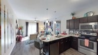 Located at 1680 Hero Way in Leander, Texas, this single-floor residence offers a thoughtfully designed open-concept layout that seamlessly connects the kitchen and living areas. The kitchen is appointed with rich dark cabinetry, granite countertops, a deep double-basin sink, and a mosaic tile backsplash that adds texture and visual interest. A peninsula with bar seating and stylish pendant lighting serves as a natural gathering point, making it ideal for casual dining or entertaining. Stainless steel appliances complement the contemporary aesthetic throughout. The living area flows naturally from the kitchen and is bathed in natural light thanks to large windows dressed with floor-to-ceiling drapes. A ceiling fan overhead adds comfort and practicality to the space. Consistent hardwood-style flooring runs throughout the open area, lending warmth and a cohesive feel. The overall ambiance is modern and inviting, with neutral wall tones providing a versatile backdrop that accommodates a variety of interior styles. The open floor plan enhances the sense of spaciousness, making this single-level residence a functional and stylish space in the heart of Leander, TX.
