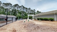 Located at 2222 Crow Ln in Myrtle Beach, South Carolina, this single-story property features a well-appointed outdoor dog park and amenity area designed for pets and their owners. The space is enclosed by a sturdy black iron fence, providing a secure environment for animals to roam freely. A charming white pergola serves as a focal point near the entrance, adding a decorative architectural touch while offering a shaded gathering spot. The ground is covered with a layer of wood chip mulch, creating a comfortable and low-maintenance surface throughout the area. Agility-style pet features and green accessory stations are thoughtfully placed across the space, enhancing its functionality. A bench provides a resting spot for visitors. The area is bordered by the exterior wall of an adjacent gray-sided building, while the backdrop is defined by a dense canopy of tall pine trees that lend a sense of privacy and natural beauty. Well-maintained shrubs line the building's edge, adding greenery and polish to the overall setting. The open layout and natural surroundings create a welcoming, park-like atmosphere ideal for outdoor recreation and pet activities.
