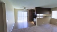 This single-floor space showcases an open-concept layout that integrates the kitchen and living area into one cohesive environment. The kitchen is equipped with dark espresso-toned cabinetry that extends to the upper wall, providing generous storage capacity. A peninsula with granite-style countertops doubles as a casual dining or workspace, while a black refrigerator and a sink with a window above complete the functional cooking area. The transition from tile flooring in the kitchen to wood-style flooring and then plush carpet in the living area subtly defines each zone while maintaining visual continuity. The living space is generously proportioned, anchored by a ceiling fan with a light fixture that provides both air circulation and ambient lighting. A large sliding glass door dressed with vertical blinds floods the interior with natural light and provides direct access to an outdoor area. The textured ceiling adds a classic residential touch throughout. Neutral wall tones complement the contrast of dark cabinetry, creating a balanced and welcoming atmosphere throughout the space.