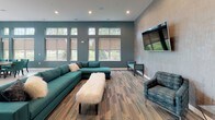 Located in Menomonee Falls, Wisconsin, this single-floor community clubhouse showcases a thoughtfully designed interior defined by a bold teal and gray color palette carried consistently throughout every zone. The open-concept layout flows seamlessly from one area to the next, creating a welcoming and functional gathering space.

The lounge area anchors the space with a generous teal sectional sofa, coordinating accent chairs, and a plush upholstered bench, all resting on patterned multi-tone carpeting. A large wall-mounted flat-screen television and high clerestory windows allow abundant natural light to pour in while offering views of the surrounding greenery.

The fully equipped kitchen is a visual centerpiece, featuring a striking oceanic blue island countertop that draws the eye immediately. White cabinetry, stainless steel appliances including a full-size refrigerator and built-in oven, and a vivid teal tile backsplash create a refined yet functional culinary space. Pendant lighting adds warmth overhead.

Adjacent to the kitchen, a formal dining area accommodates a large wooden table surrounded by tufted teal upholstered chairs. An industrial-style multi-globe chandelier hangs above, while oversized windows frame outdoor views. A secondary lounge nook with patterned seating and a wall-mounted television completes the space, offering a more intimate setting for smaller gatherings.