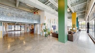 Located at 300 18th St S in Birmingham, Alabama, this dynamic single-floor commercial space seamlessly blends industrial architecture with bold, contemporary interior design. The entry welcomes visitors through sleek floor-to-ceiling glass curtain walls framed in black steel, flooding the interior with abundant natural light and offering open views of the surrounding streetscape.

Throughout the space, polished concrete floors provide a clean, durable foundation that complements the exposed ceiling structure, complete with visible ductwork, textured spray insulation, and industrial lighting. Signature colorful dash-pattern murals in teal, orange, gold, and green animate the white walls, lending a cohesive and energetic visual identity across all areas.

The main lounge area features a generous seating arrangement with plush leather sofas, accent chairs, layered rugs, and curated decorative shelving — ideal for collaborative or social gatherings. A dedicated meeting and work zone offers table seating alongside wall-mounted displays, supporting productive work sessions. An additional lounge nook showcases eclectic furnishings, a striking woven wall hanging, and open shelving with artful accessories.

Bold structural columns painted in green and yellow punctuate the open floor plan, adding architectural character. The overall ambiance is creative, welcoming, and distinctly curated — well-suited for a coworking, event, or creative studio environment.