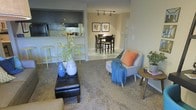 This single-floor space is designed with an open-concept layout that seamlessly connects the living area, dining space, and kitchen. The living area features plush carpeted flooring and is furnished with a sleek black leather ottoman serving as a coffee table, complemented by a light grey accent chair adorned with an orange pillow and blue throw blanket. Decorative vases and greenery add a touch of natural warmth to the space.

Transitioning into the dining area, the flooring shifts to tile, providing a clean and practical surface underfoot. A dark wood dining table surrounded by matching chairs sits beneath track lighting mounted on the ceiling, offering focused illumination over the space. Framed artwork on the neutral-toned walls adds a curated, finished look to the area.

The kitchen is accessible via a pass-through counter that doubles as a breakfast bar, fitted with tall bar stools in a bright yellow-green hue, adding a pop of color. The kitchen features dark cabinetry and overhead appliances, providing a functional and stylish cooking space. Overall, the layout is efficient and cohesive, with a warm, inviting ambiance throughout.