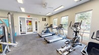 Located at 1404 Meads Rd in Norfolk, Virginia, this fitness center offers a thoughtfully designed, single-floor workout facility with a generous open floor plan. The space is bathed in natural light streaming through multiple windows positioned along the walls, providing pleasant outdoor views of the surrounding greenery. Recessed fluorescent lighting and ceiling fans maintain a bright, well-ventilated environment throughout.

The facility is divided into distinct workout zones. The cardio area features a row of treadmills, elliptical trainers, and a stationary bike, all positioned to take advantage of the natural light and outdoor views. The strength-training zone is outfitted with a multi-station cable machine, a leg press, and additional resistance equipment, offering a comprehensive range of muscle-targeting options. A dedicated free weights area includes a full dumbbell rack, adjustable weight benches, medicine balls, and a large wall mirror that enhances the sense of space.

Motivational wall art adds personality to the space, while the neutral-toned walls and durable carpeted flooring create a clean, functional aesthetic. A glass-panel entryway connects to an adjacent area, and direct exterior access adds convenience. The overall ambiance is welcoming and purposeful, making this fitness center a fully equipped destination for a complete workout experience.