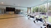This single-floor fitness facility is thoughtfully designed to support a wide range of workout disciplines within a clean, contemporary setting. The space is anchored by a spacious open-floor training zone finished with light hardwood flooring, providing an inviting and functional surface ideal for group classes, stretching, or bodyweight exercises. A dedicated Training Zone wall rack is stocked with an extensive selection of equipment, including kettlebells, medicine balls, TRX suspension trainers, and weighted plates, offering everything needed for strength and functional training.

A full-length mirror wall visually expands the main workout area, enhancing both spatial perception and training utility. Glass partition walls with sleek aluminum framing divide the space into distinct zones while maintaining an open, connected feel throughout. The spin studio is a standout feature, lined with professional-grade spinning bikes and framed by expansive floor-to-ceiling windows that overlook a lush, green outdoor landscape, flooding the room with natural light and creating an energizing atmosphere.

Recessed lighting throughout ensures consistent, well-distributed illumination across all areas. The overall design balances functionality with a polished, modern aesthetic, making this a well-equipped and visually appealing fitness environment.