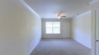 This single-floor residential space presents a straightforward and functional layout designed for comfortable everyday living. The main living area is finished with wall-to-wall carpet in a neutral tone, providing a soft and inviting surface underfoot. Crown molding lines the upper perimeter of the room, adding a subtle architectural detail that elevates the overall finish of the space. A ceiling fan with an integrated light fixture is centrally positioned, offering both air circulation and ambient lighting throughout the room. A window framed with a classic grid-pane design allows natural light to flow into the space, brightening the interior and creating a pleasant, airy atmosphere. Adjacent to the window, a solid white entry door provides access to the exterior. The walls are finished in a clean, light neutral tone that complements the bright and open feel of the room. Baseboards run consistently along the lower walls, contributing to the tidy, well-maintained character of the space. Overall, the unit offers a clean, move-in ready environment with classic residential finishes suited for practical, day-to-day living.
